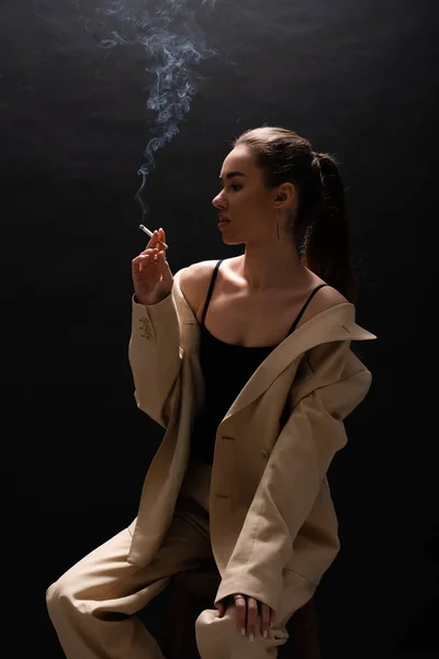 Young brunette woman in beige blazer holding cigarette while sitting on black background — Stock Photo