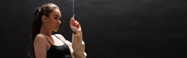Young brunette woman in beige blazer holding cigarette while looking away on black background, banner — Stock Photo
