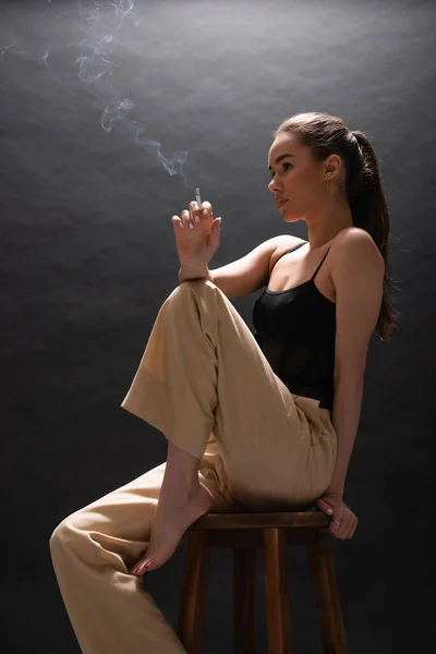 Босоногая женщина в бежевых штанах, держащая сигарету, сидя на высоком стуле на черном фоне — стоковое фото