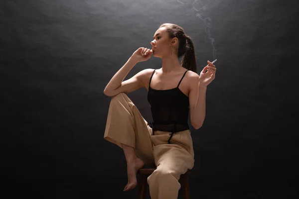 Thoughtful woman in beige pants holding cigarette while sitting on high chair on black background — Stockfoto