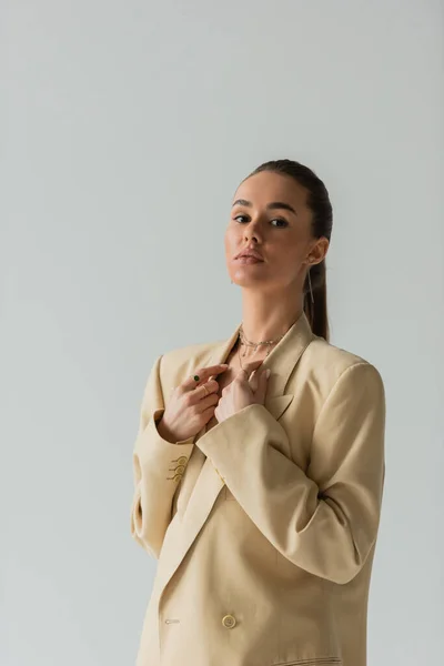 Brunette young woman in beige jacket posing while looking at camera isolated on grey — Stockfoto