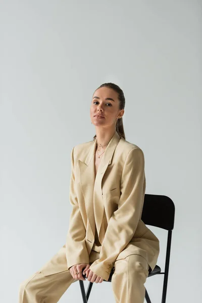 Brünette junge Frau im beigen trendigen Anzug sitzt auf Stuhl isoliert auf grau — Stockfoto
