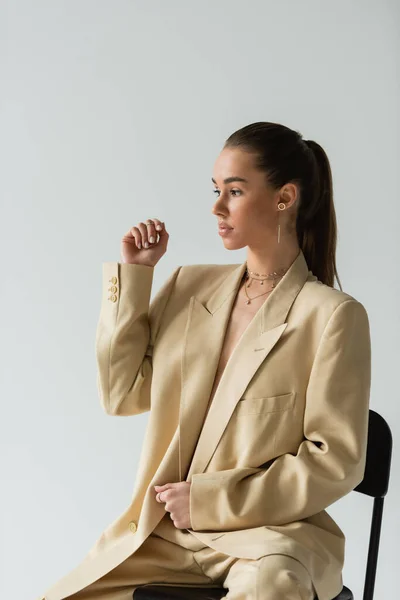 Pretty young woman in beige trendy suit sitting on chair isolated on grey — Fotografia de Stock