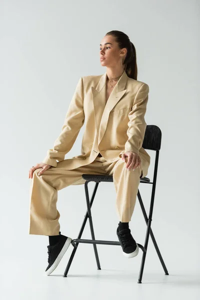 Brünette junge Frau im beigen trendigen Anzug sitzt auf Stuhl auf grau — Stockfoto
