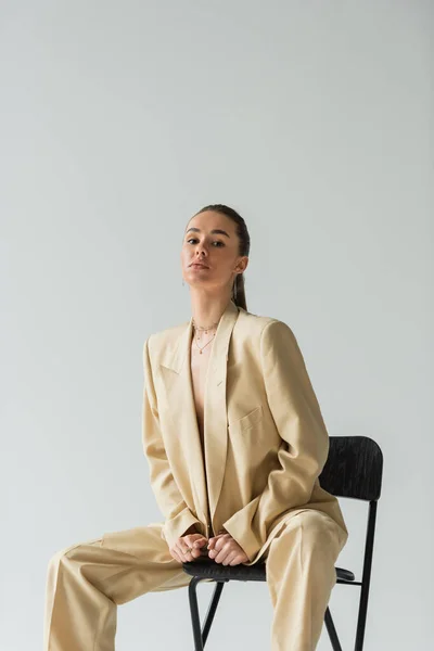 Брюнетка жінка в модному і бежевому костюмі сидить на стільці ізольовано на сірому — стокове фото