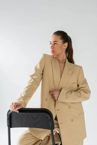 Pretty young woman in beige jacket leaning on chair back while posing isolated on grey — Stock Photo