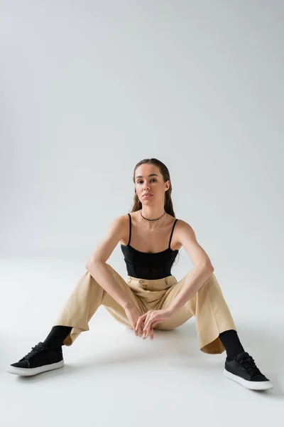 Full length of pretty model in beige pants and black top looking at camera while sitting on grey — Stock Photo