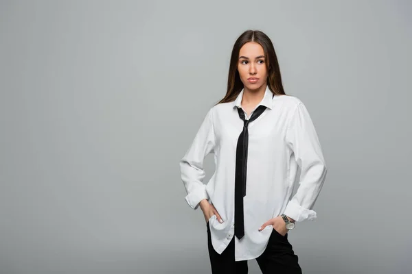 Young and brunette woman in white shirt with tie posing with hands in pockets isolated on gray — Stock Photo