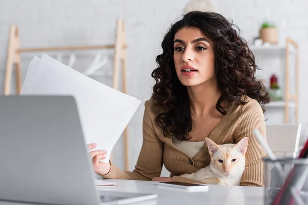 Morena copywriter segurando papéis e gato oriental perto de laptop em casa — Fotografia de Stock