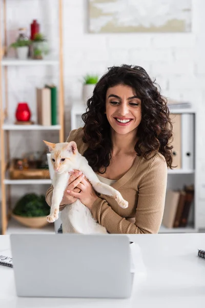 Smiling copywriter holding oriental cat near laptop at home — Stock Photo
