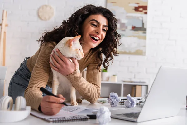 Happy copywriter petting oriental cat near papers and laptop at home — Stock Photo
