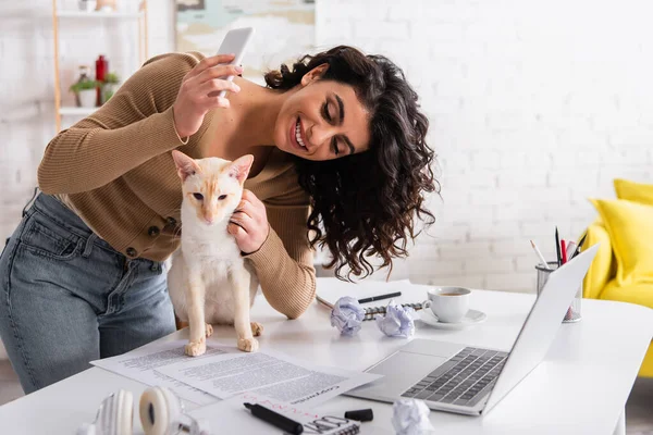 Cheerful copywriter holding smartphone and petting oriental cat near papers and laptop — Photo de stock