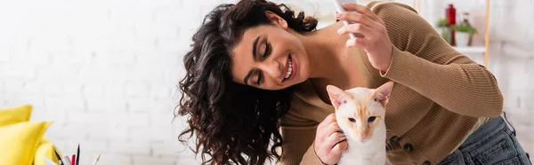 Mujer sonriente sosteniendo teléfono celular y acariciando gato oriental en casa, pancarta — Stock Photo