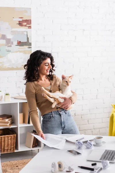 Brünette Werbetexterin hält orientalische Katze in der Nähe von Dokumenten und Gadgets zu Hause — Stockfoto