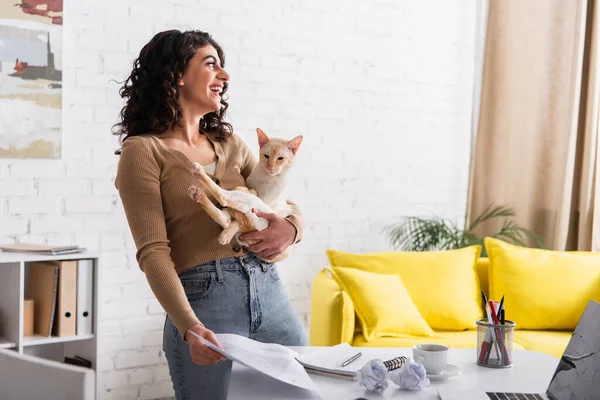 Side view of cheerful copywriter holding oriental cat and papers at home — Photo de stock