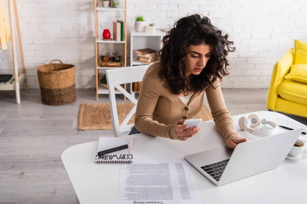 Curly copywriter using gadgets near papers and headphones on table at home — Stock Photo