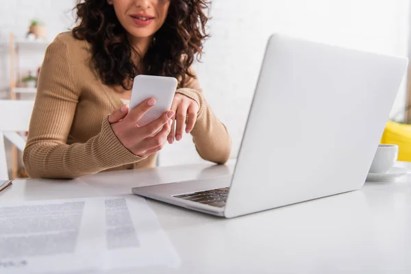 Cropped view of copywriter using smartphone near laptop and blurred papers at home — Stock Photo