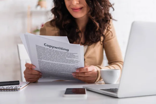 Cropped view of smiling woman holding papers with copywriting lettering near gadgets and coffee at home — Stock Photo