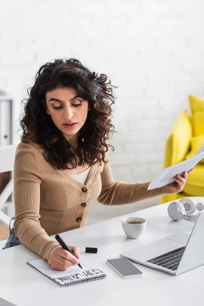 Curly copywriter holding papers and writing on notebook near devices on table at home — Stock Photo