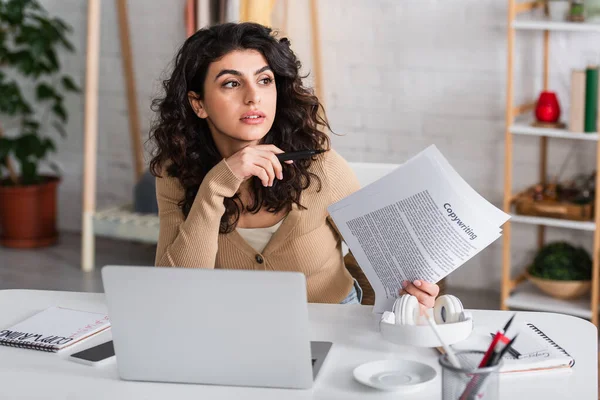 Brunette copywriter holding papers near gadgets and headphones in living room — Stock Photo