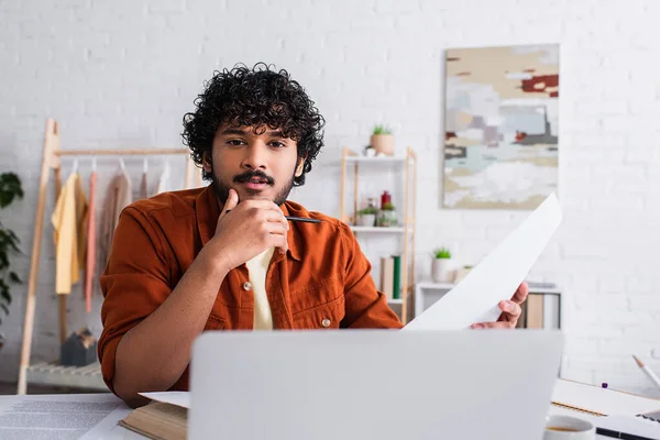 Young indian copywriter holding paper and looking at camera near blurred laptop at home — Stock Photo
