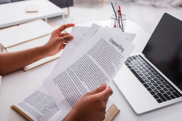 Обрезанный вид фрилансера с бумагами с копирайтингом рядом с книгой и ноутбуком дома — стоковое фото