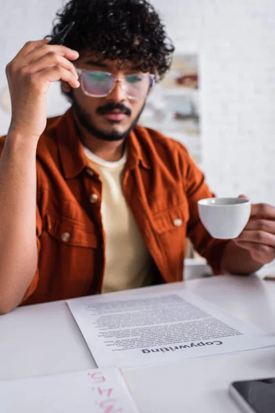 Blurred indian copywriter holding cup near paper and cellphone on table — Stock Photo