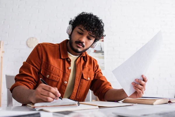 Indian copywriter in headphones holding papers and writing on notebook at home — Photo de stock