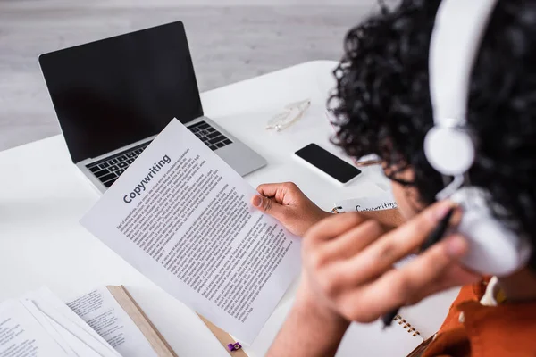 Devices near blurred indian freelancer in headphones working with paper with copywriting lettering at home — Photo de stock