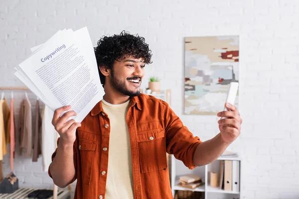 Smiling indian copywriter having video call on smartphone and holding papers at home — Stock Photo