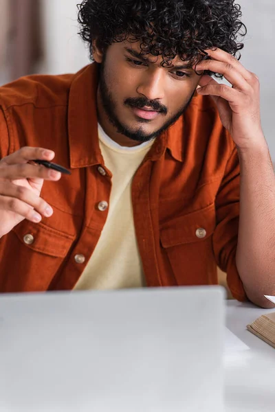 Focused indian copywriter holding pen near blurred laptop at home — Stock Photo