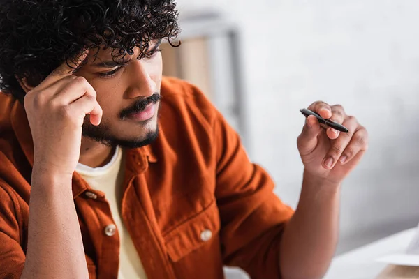 Focused indian copywriter holding pen near blurred book at home — Stock Photo