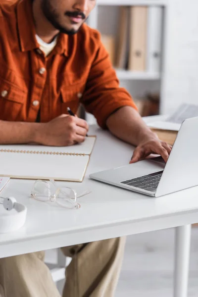 Cropped view of indian copywriter using laptop and writing on notebook near eyeglasses at home — Stock Photo