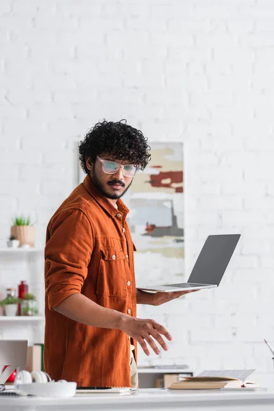 Indian copywriter in eyeglasses holding laptop near book and headphones at home — Stock Photo
