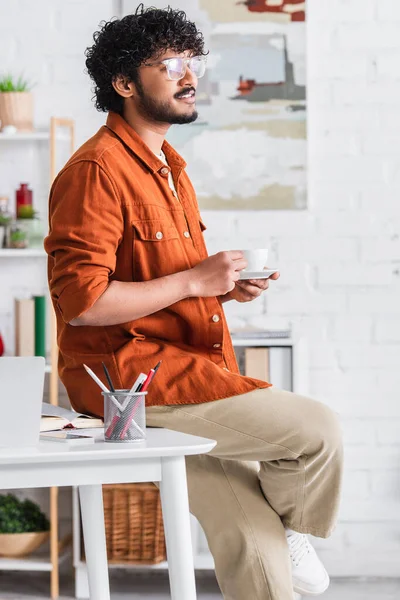 Smiling indian copywriter holding coffee near devices and book at home — Stock Photo