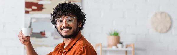 Smiling indian man in eyeglasses holding cup of coffee at home, banner — Stock Photo