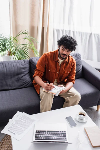 Indian copywriter writing on notebook near devices in living room — Stock Photo
