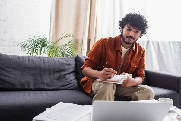 Indian copywriter writing on notebook near laptop and coffee in living room — Stock Photo