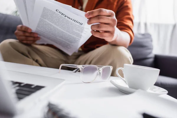 Cropped view of freelancer holding papers with copywriting lettering near coffee and eyeglasses at home — Stock Photo