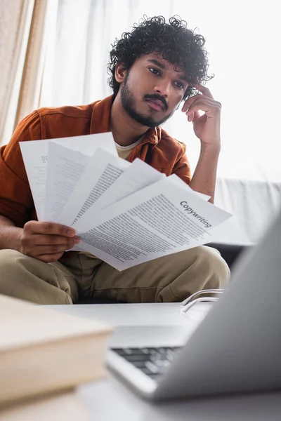 Concentrated indian freelancer holding papers with copywriting lettering near blurred laptop in living room — Stock Photo
