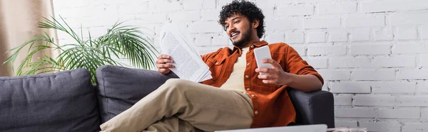Cheerful indian copywriter looking at papers and holding smartphone on couch at home, banner — Stock Photo