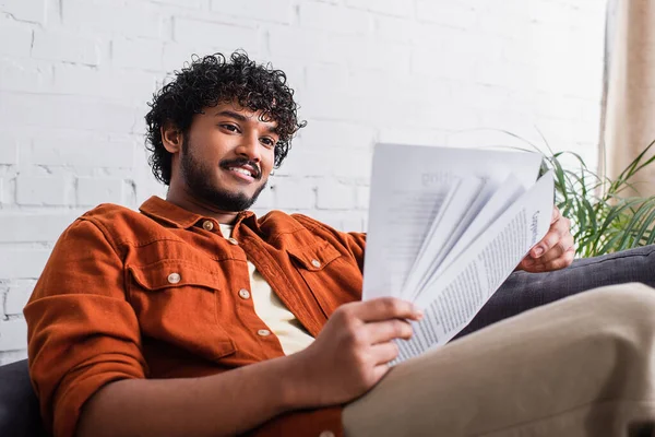 Positive indian copywriter looking at papers while sitting on couch at home — Fotografia de Stock