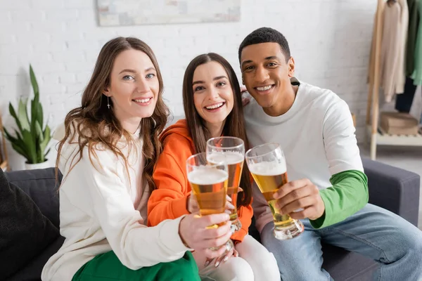 Cheerful women and african american man clinking glasses of beer while looking at camera in living room — Stock Photo