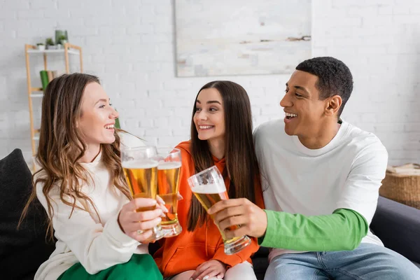 Mujeres alegres y sonriente hombre afroamericano tintineo vasos de cerveza en la sala de estar — Stock Photo