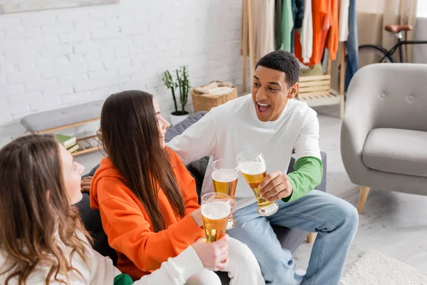 Веселі жінки і здивований афроамериканський чоловік кляне келихи пива у вітальні — стокове фото