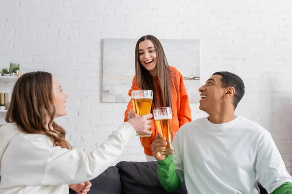 Mujeres positivas y sorprendidos afroamericanos hombres tintineo vasos de cerveza en la sala de estar - foto de stock