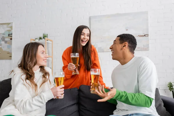 Mulheres positivas e homem americano africano feliz segurando copos de cerveja na sala de estar — Fotografia de Stock