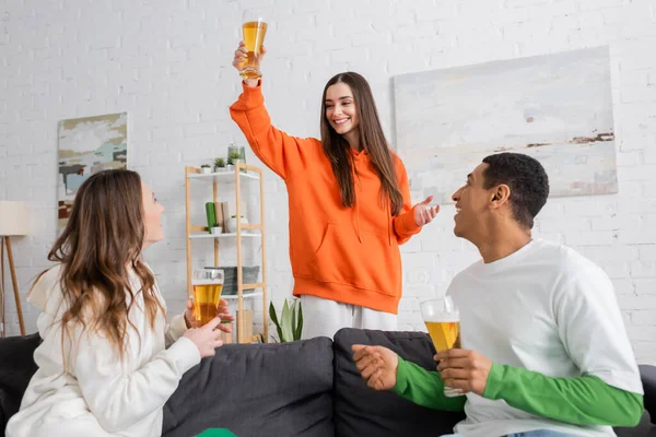 Glückliche Frau prostet Glas Bier neben interrassischen Freunden im Wohnzimmer zu — Stockfoto