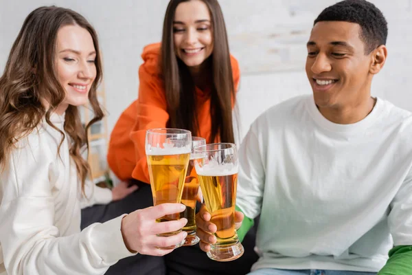 Positive women and cheerful african american man toasting glasses of beer in living room — Stock Photo