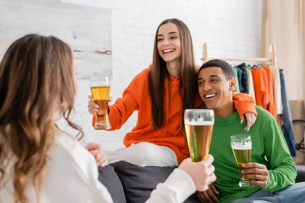 Femme heureuse étreignant homme afro-américain joyeux tout en tenant un verre de bière et en regardant ami — Photo de stock